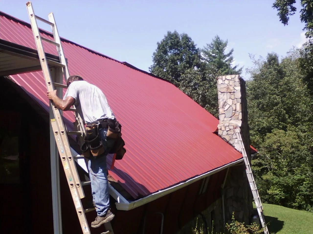 Roofer on a ladder beside a red metal roof and stone chimney — Soffit & Fascia Repair by Roofing Brothers in Mc Dermitt